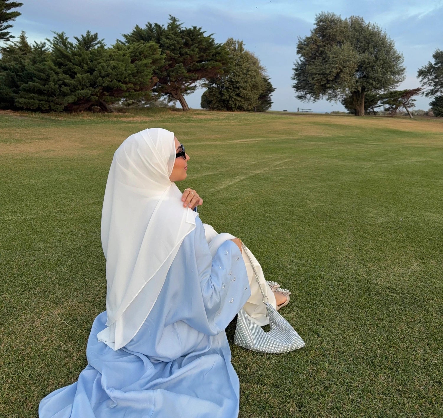 Woman in a light blue abaya with crescent moon embroidery and white hijab, sitting on green grass in an Australian park at golden hour, holding a sparkly handbag