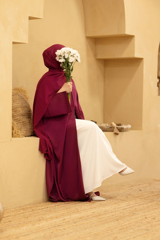 Woman wearing a burgundy classic abaya with matching hijab, sitting indoors while holding flowers — Classic Abaya Burgundy Bliss.