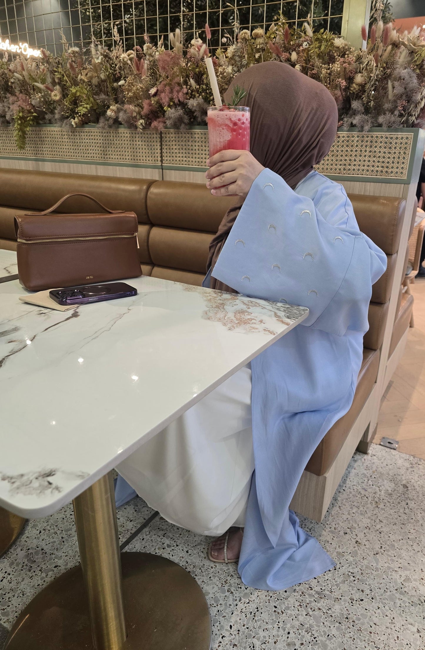 Person seated at a café table, enjoying a pink drink with a straw while wearing a light blue abaya.