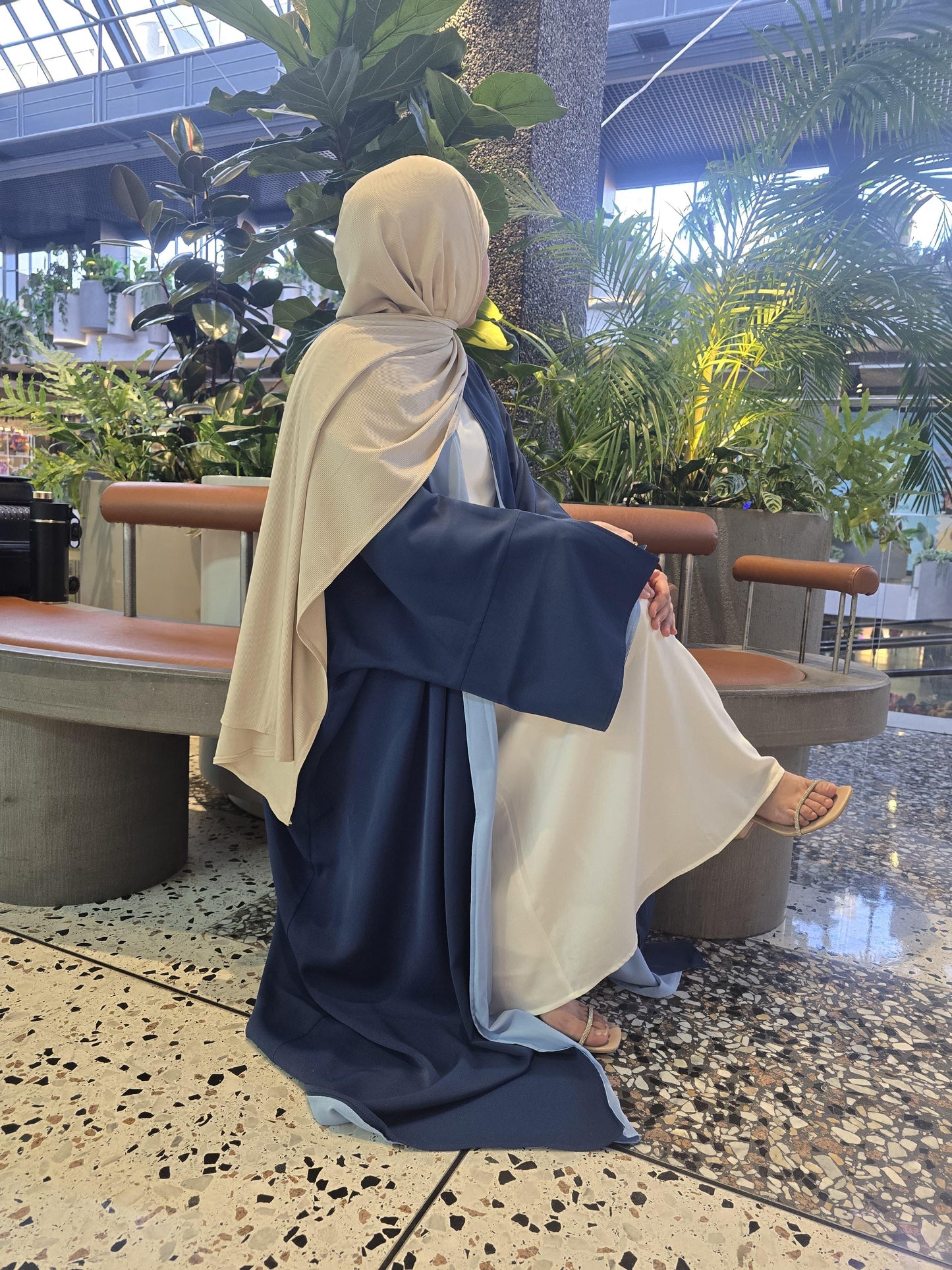 Woman sitting on a bench wearing Reversible Abaya Midnight Sky and beige hijab, taking a selfie indoors with plants around.
