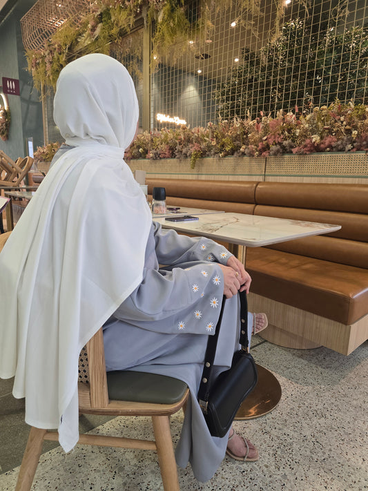 A woman wearing the Zahra Abaya Silver Mist, sitting indoors on a chair with a soft, elegant background.